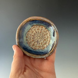 May include: A small, round ceramic dish with a mandala design. The dish features a raised, detailed mandala in the center, surrounded by a blue and brown glazed rim. The dish is held in a hand.