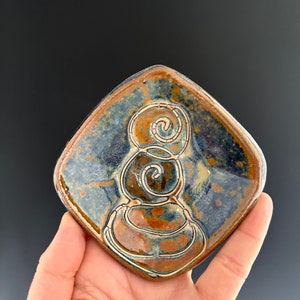 May include: A square ceramic dish with a blue and brown glaze. The dish has a raised design of a spiral with three circles.