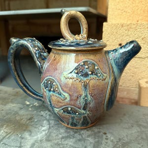 May include: A handcrafted ceramic teapot with a lid and handle. The teapot features a mushroom design in shades of brown, blue, and purple. The handle and spout are a dark blue color. The teapot is sitting on a gray surface.