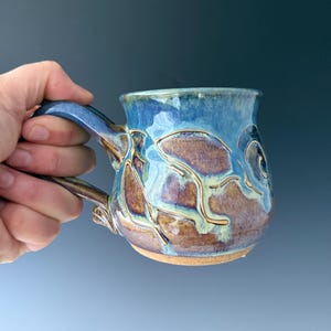 May include: A handmade ceramic mug with a blue glaze and brown accents. The mug features a unique design with raised patterns and a curved handle. The mug is held by a hand, showcasing its size and shape. The mug is approximately 4 inches tall.