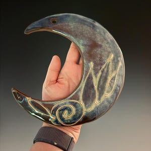 May include: A crescent moon-shaped ceramic wall hanging with a glossy, variegated glaze in shades of deep blue, purple, and brown. The surface features a carved, light-colored floral design with swirling lines and petals etched into the clay.