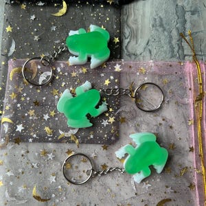 May include: Three green and white frog-shaped keychains with silver key rings. The keychains are displayed on a surface with two small drawstring bags. One bag is black with gold stars and moons, and the other is pink with gold stars.