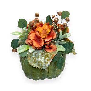 May include: A green pumpkin with a floral arrangement of orange hydrangeas and gold berries. The pumpkin is decorated with green leaves and has a speckled texture.