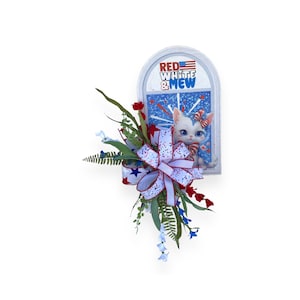 May include: A white cat with red and blue bow peeks out of a window with a blue and white background. The window is framed in white and has the text "Red White & Mew" in red and blue letters. The window is decorated with a red, white, and blue bow and greenery.