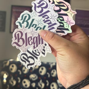 以下が含まれることがあります： ゴシック調のフォントで「Bleah」という文字がデザインされた、ダイカットステッカーのコレクション。各ステッカーには小さな星のアクセントが施されており、紫、ティール、虹色のグラデーションなど、それぞれ異なるカラーバリエーションで構成されています。