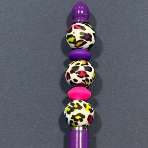 May include: A purple pen with a decorative top. The top features a series of colorful beads, including leopard print spheres, a pink oval, and a purple finial. The pen's body is also purple.