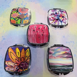 May include: Five compact mirrors with different colorful designs. The mirrors are square shaped with silver frames. The designs include a pink flamingo pattern, a colorful abstract design, a yellow flower design, a pink and blue sunset design, and a floral design with green leaves.