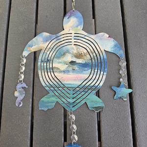 May include: A metal sea turtle wind spinner featuring a blue, white, and pink painted ocean scene. The turtle has concentric cutouts and is decorated with three hanging chains of clear beads, ending in a seahorse, a starfish, and a seashell charm.