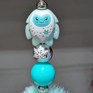 May include: A whimsical keychain featuring a light blue yeti charm with a snowflake design, silver beads, a turquoise ball, and a fluffy, light blue pom-pom. The keychain is attached to a green clasp.