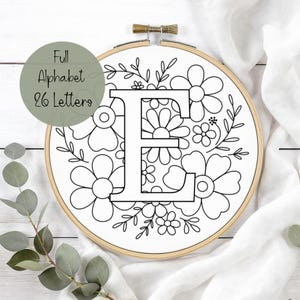 Puede incluir: Un bastidor de bordado con un diseño floral y la letra "E" en el centro. El diseño es en blanco y negro, con el texto "Full Alphabet 26 Letters" en un círculo verde. El bastidor es de madera.