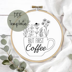 May include: A white embroidery hoop with a floral design and the text "BUT FIRST Coffee." The design features a coffee cup filled with line art flowers and foliage. A PDF template label is in the upper left corner. The hoop is made of wood.