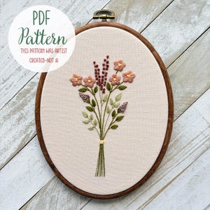 Op de afbeelding: Een ovale borduurring met een bloemenontwerp. De borduursel toont een boeket van roze en paarse bloemen met groene bladeren, vastgebonden met een gele draad. De tekst "PDF Pattern" en "THIS PATTERN WAS ARTIST CREATED-NOT AI" staat in een witte cirkel.
