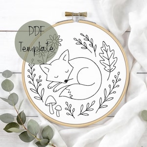 Puede incluir: Un bastidor de bordado de tela blanca con un diseño de zorro durmiente, rodeado de hojas, flores y setas. El texto "PDF Template" está superpuesto. El diseño está en un bastidor de madera.