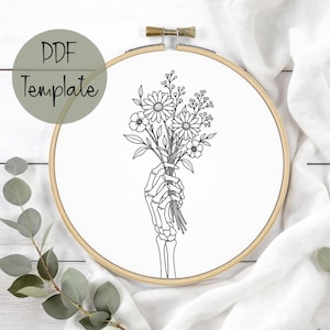 May include: A circular wooden embroidery hoop containing white fabric with a black line art illustration of a skeletal hand holding a bouquet of wildflowers. The text PDF Template is printed in the upper left corner of the image.