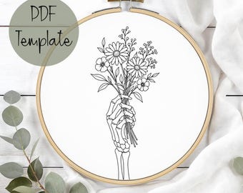 May include: A circular wooden embroidery hoop containing white fabric with a black line art illustration of a skeletal hand holding a bouquet of wildflowers. The text PDF Template is printed in the upper left corner of the image.