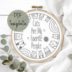 May include: A PDF template for an embroidery hoop art project. The design features the text "Cats Are My Favorite People" surrounded by illustrated cats, flowers, and a rainbow. The hoop is made of wood with a brass-colored clasp.
