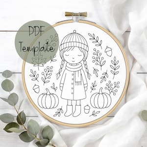 以下が含まれることがあります： 冬の服装の女の子の白黒線画が刺繍枠に描かれています。秋をテーマにした要素に囲まれています。「PDF Template」というテキストが緑色の円の中にあります。デザインにはカボチャ、葉、ドングリが含まれています。