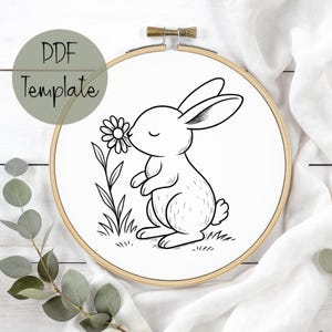 May include: An embroidery hoop featuring a black and white illustration of a rabbit smelling a flower. The text "PDF Template" is in a green circle. The design is on white fabric, within a wooden hoop.
