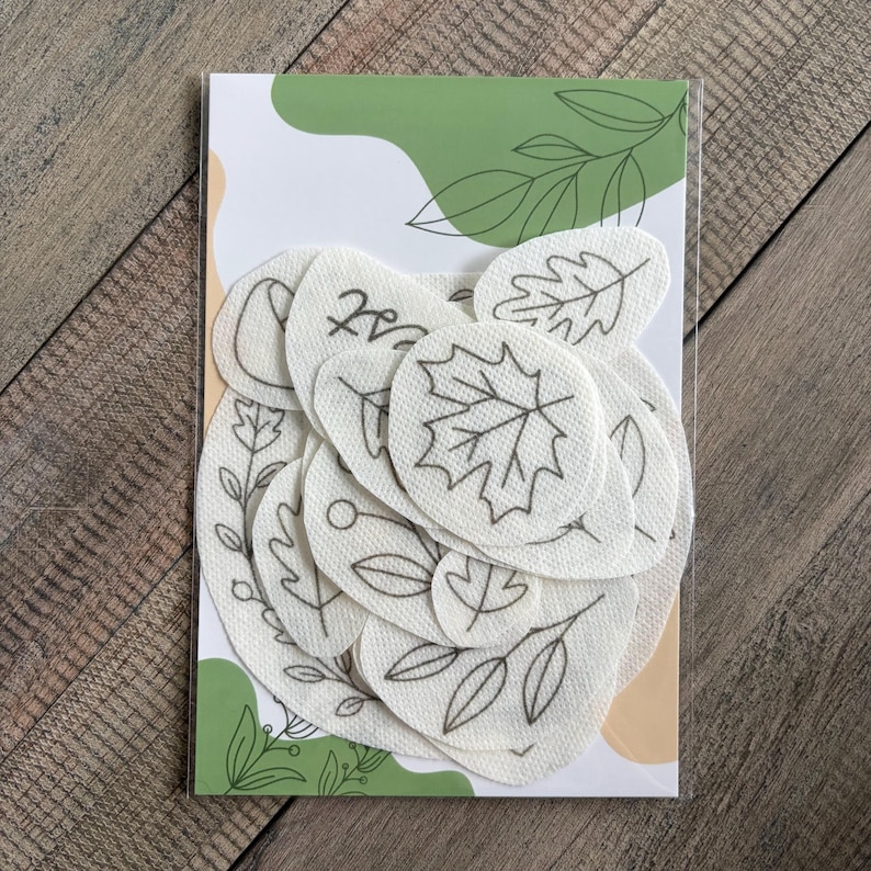 May include: A collection of off-white fabric patches featuring black line art illustrations of various leaves, branches, and berries. The patches are arranged in a stack on a rectangular backing card decorated with green and tan abstract shapes.