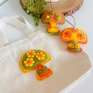May include: Three felt mushroom ornaments with floral details in shades of orange, yellow, and green. The mushrooms are placed on a white tote bag and a wooden slice. Each mushroom has a brown string for hanging.