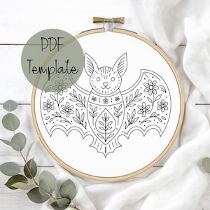 May include: A black and white bat embroidery design in a wooden hoop. The bat is decorated with floral and leaf patterns. The text "PDF Template" is in a green circle in the upper left corner. The background is white fabric.