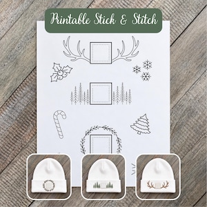May include: A white sheet with black line art designs including antlers, holly, snowflakes, pine trees, a candy cane, and a wreath, each framing a square. Below are three white beanies featuring embroidered versions of these designs.