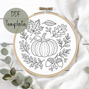May include: A white fabric stretched within a wooden embroidery hoop, featuring a black line drawing of a pumpkin, leaves, acorns, and berries. The text "PDF Template" is in a green circle. The design evokes autumn.