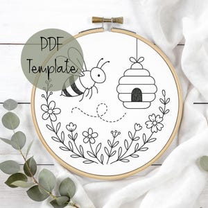 Puede incluir: Un tejido blanco dentro de un bastidor de bordado de madera, con un dibujo lineal en blanco y negro de una abeja, una colmena y elementos florales. El texto "PDF Template" está superpuesto en la imagen.