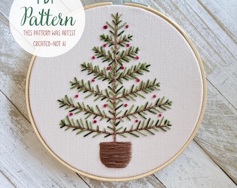 May include: An embroidery hoop containing a white fabric panel with a hand-stitched Christmas tree design. The tree features green branches, small pink berry accents, and a brown trunk in a pot. Text reads: PDF Pattern, This pattern was artist created-not AI.