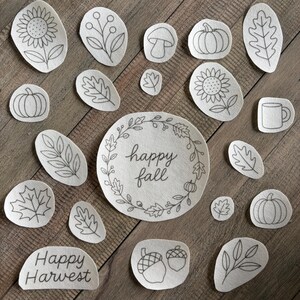 May include: A collection of off-white fabric patches featuring black line art illustrations of autumn themes including pumpkins, sunflowers, leaves, acorns, a mushroom, a mug, and text reading Happy Fall and Happy Harvest in various shapes.