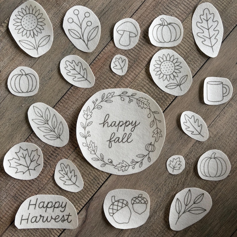 May include: A collection of off-white fabric patches featuring black line art illustrations of autumn themes including pumpkins, sunflowers, leaves, acorns, a mushroom, a mug, and text reading Happy Fall and Happy Harvest in various shapes.