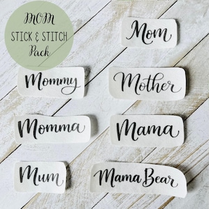 May include: White fabric labels with black cursive text, including "Mom," "Mommy," "Mother," "Momma," "Mama," "Mum," and "Mama Bear." The labels are arranged on a white wooden surface. The top left corner has a green circle with the text "Mom Stick & Stitch Pack."