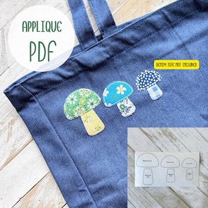 Puede incluir: Un bolso tote de mezclilla azul con tres apliques de setas en varios patrones y colores. Los apliques incluyen una seta verde con tallo amarillo, una seta azul con una flor blanca y una seta de lunares azules y blancos. El texto "APPLIQUE PDF" está en un círculo.