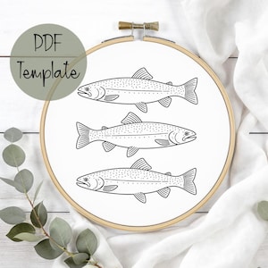 Puede incluir: Un bastidor de bordado con tela blanca que muestra tres ilustraciones de peces en blanco y negro. El texto "PDF Template" está en un círculo verde. Adecuado para proyectos de bordado o punto de cruz.