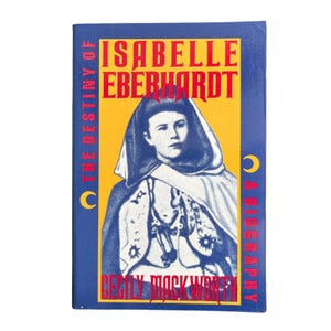 May include: A hardcover book titled "Isabelle Eberhardt: A Biography" by Cecily Mackworth. The cover features a portrait of a woman in a hooded cloak against a yellow background, with text in red and blue.