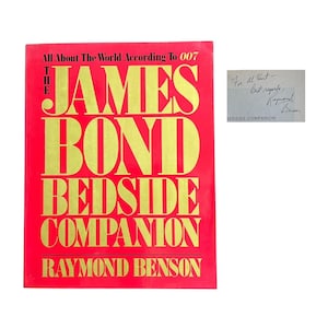 May include: A red book titled "The James Bond Bedside Companion" with gold lettering. The author's name, Raymond Benson, is at the bottom. A handwritten note is included, with the words "For Al Haut - Best regards, Raymond Benson."