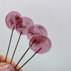 May include: Four pink lollipop sticks with gold glitter embedded in the candy. The lollipops are held in a hand.