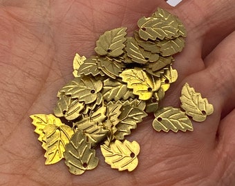 May include: A pile of gold-colored metal leaf charms with two small holes in each leaf. The leaves are arranged in a random pattern and are all the same size and shape.