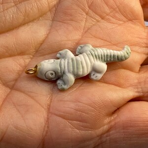 May include: A small, white ceramic lizard charm with a gold loop for hanging. The lizard has a detailed, textured body and large, black eyes.