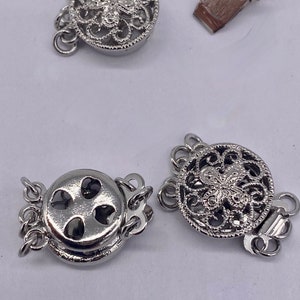 Puede incluir: Dos cierres de metal plateados para collares o pulseras. Un cierre tiene un diseño de filigrana decorativo con una mariposa en el centro. El otro cierre tiene un diseño simple con tres recortes en forma de corazón.