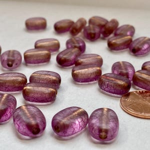 May include: A collection of oval-shaped beads in shades of purple and bronze. The beads have a crackled texture and are scattered across a surface. A penny is included for size reference.
