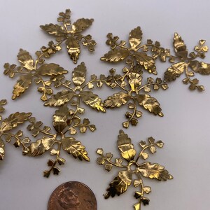 May include: A group of eight gold metal leaf-shaped embellishments with a hole in the center. The embellishments are arranged in a random pattern on a white surface. A penny is included for scale.