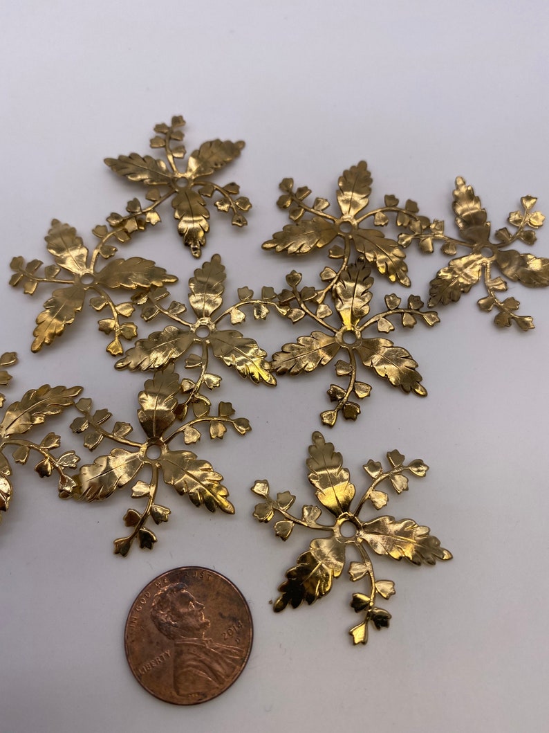 May include: A group of eight gold metal leaf-shaped embellishments with a hole in the center. The embellishments are arranged in a random pattern on a white surface. A penny is included for scale.