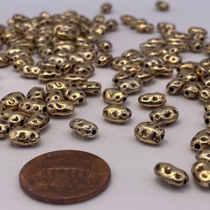 May include: A pile of gold-toned metal beads with a textured surface. The beads are oval-shaped and have a hole through the center. A penny is included for scale.