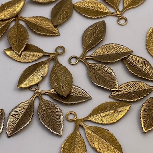 Peut inclure: Un groupe de 10 breloques en forme de feuilles en métal doré avec une boucle en haut. Chaque breloque a trois feuilles avec une surface texturée.