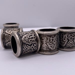 Pode incluir: Quatro contas de cabelo de metal prateadas com um design floral intrincado. As contas são redondas e têm um acabamento liso e polido.