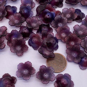 Könnte beinhalten: Eine Sammlung von violetten, irisierenden Plastikblumenverzierungen. Die Blumen haben fünf Blütenblätter und ein kleines Loch in der Mitte. Die Blumen sind auf einer weißen Oberfläche mit einem Penny als Größenvergleich verstreut.