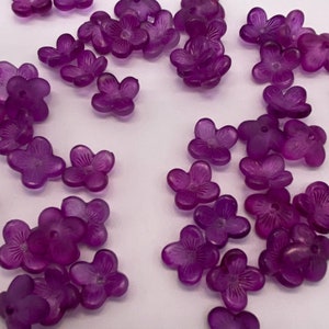 May include: A collection of purple plastic flower-shaped beads. The beads are scattered on a white surface.