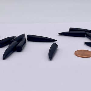 May include: A group of black, pointed, horn-shaped beads, some with a hole drilled through the top. The beads are scattered on a white surface, with a penny for scale.