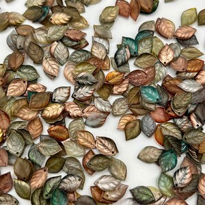 May include: A collection of leaf-shaped beads in various metallic colors, including copper, bronze, silver, and green. The beads have a textured surface and a small hole at the top for stringing. The leaves are scattered across a white surface.